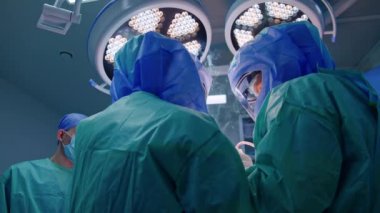 Doktorların arka görüşü duman çıkaran elektrikli bir cihaz kullanıyor. Koruyucu miğferli sıhhiyeler yuvarlak lambalar altında çalışır. Düşük açılı perspektif.