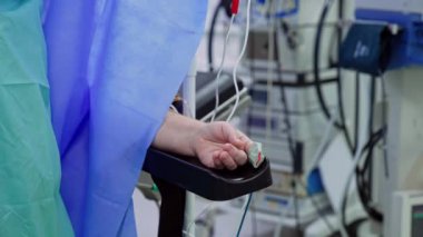 İşaret parmağında sensör olan bir hastanın eli. Mavi bornozlu tanınmayan doktor operasyon masasına geliyor..