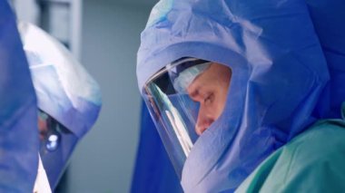 Koruyucu kasklı beyaz erkek doktor aşağıya bakıyor. Ameliyatta meşgul bir cerrahın yan görünüm portresi.