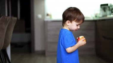 Mutfağın yanından yürüyüp elma yiyen küçük çocuk. Bebek oturur ve meyveyi yere koyar..