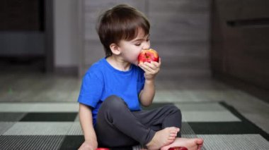 Güzel erkek bebek büyük kırmızı bir elma yiyor. Çocuk önünde elmalarla mutfak zemininde oturuyor..