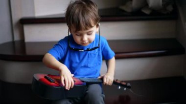 Merdivenlerde oturan, güneş gözlüğü takan ve küçük gitar çalan sevimli küçük çocuk. Evde oynayan sevimli çocuk..
