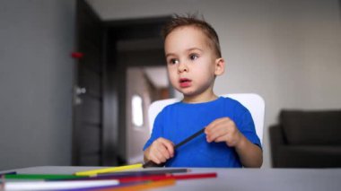 Mavi tişörtlü sevimli erkek bebek elinde kalemle masada oturuyor. Çocuk kalemi koyar ve ayağa kalkar. Düşük açı görünümü.