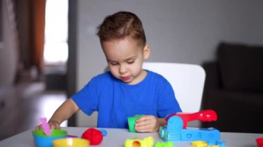 Mavi tişört giyen güzel, beyaz bir bebek masada oturuyor. Çocuk bir sopayla plastikini kesmeye çalışıyor..