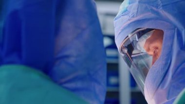 Miğferli doktor ameliyat alanına bakıyor. Ameliyat üzerine odaklanmış bir cerrahın portresi. Yan görünüm.