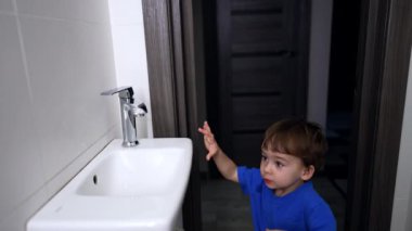 Siyah saçlı, mavi tişörtlü çocuk banyoya giriyor. Çocuk lavaboya yaklaşır ve musluğu açar..