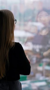 Camda duran gözlüklü çekici iş kadını. Uzun saçlı kadın şehir manzarasında pencereden dışarı bakıyor. Dikiz aynası. Dikey video