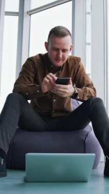 Pencere arkasında aletleri olan genç bir adam. Yakışıklı adam ofisteki koltukta oturuyor ve cep telefonu kullanıyor. Dizüstü bilgisayar yerde. Serbest meslek kavramı. Dikey video