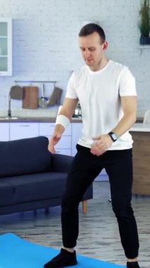Ev antrenmanı. Beyaz tişörtlü ve siyah pantolonlu genç adam mutfak içinde çömelme egzersizi yapıyor. Spor eğitimi kapalı alanlarda. Ağır çekim. Dikey video