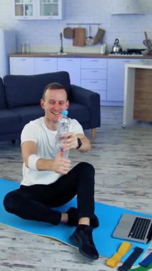 Genç adam evde egzersiz yapıyor. Adam yerde bir paspasın üzerinde oturuyor ve biberonla dengesini koruyor. Karantina sırasında evde spor yapmak. Dikey video