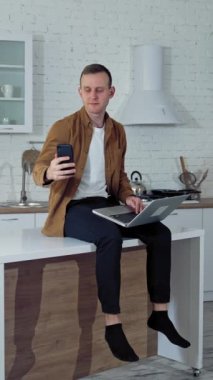 Evde online çalışan genç bir adam. Mutfak masasında dizüstü bilgisayarla oturan ve telefonla video görüşmesi yapan serbest çalışan bir erkek. Uzak bir iş. Dikey video