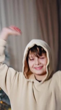 Kapüşonlu genç çocuk heyecanla atlıyor. Mutlu çocuk kapüşonunu çıkarıp saçlarını kirletiyor. Noel ağacı arka planda. Dikey video