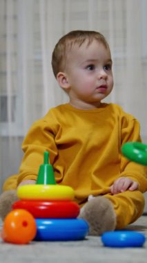 Turuncu takım elbiseli sevimli bebek oyuncaklarla çevrili oturuyor. Tatlı çocuk yana bakıyor ve çorabını ayağından çıkarıyor. Dikey video