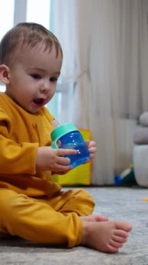 Sevimli yalınayak çocuk yerde oturmuş şişeden içiyor. Bebek şişeyi yüksekte tutuyor ve üzerine biraz su döküyor. Dikey video