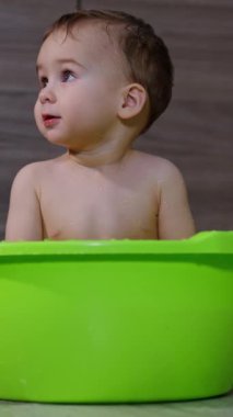 Küçük sevimli çocuk lavaboda çıplak oturuyor. Bebek etrafına şaşkın şaşkın bakıyor. İçeride yıkanma zamanı. Dikey video