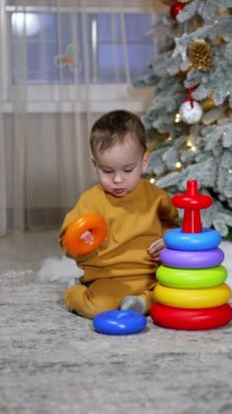 Sevimli bebek yüzükleri oyuncak piramidinden topluyor. Tatlı çocuk süslü bir Noel ağacıyla odada oynuyor. Dikey video