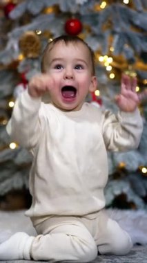Mutlu, güzel, heyecanlı çocuk ellerini sallıyor. Bebek yerde sevinçle oturuyor ve gülüyor. Noel ağacı arka planda. Dikey video