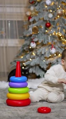 Noel ağacının yanında yerde oynayan ciddi, sakin bir bebek. Sevimli çocuk oyuncak piramidini topluyor. Dikey video