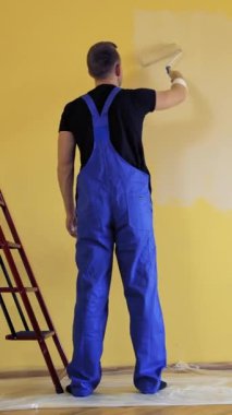 Mavi tulumlu, duvarları silindirle boyayan bir adam. İşçi dekorasyon odası yeni bir renkle. Resim hizmetleri. Dikiz aynası. Dikey video