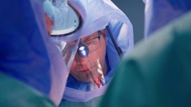 Orta yaşlı, gözlüklü, farlı ve kasklı beyaz bir erkek doktor. Operasyonda baş cerrah olarak çalışıyor. İki doktor kameraya arkasını döner. Kapat..