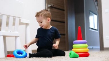 Şirin, gülümseyen, beyaz bir çocuk yerde oturuyor. Küçük çocuk piramidi odasında monte ediyor..