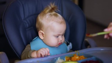 Yüksek sandalyede oturan sevimli beyaz oğlan kaşıkla besleniyor. Anne bir bebeğe püre sebze veriyor. Kapat..