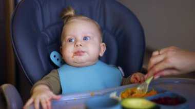 Ağzının etrafında yemek olan mavi gözlü sarışın bebek yüksek sandalyede oturuyor. Anne bir çocuğa püre sebze verir..