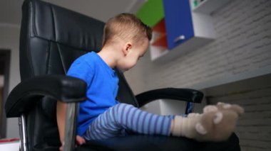 Mavi tişörtlü ve çizgili pantolonlu beyaz çocuk büyük koltukta oturuyor. Küçük çocuk bir akıllı telefon aldı ve oynamaya başladı. Düşük açı görünümü.