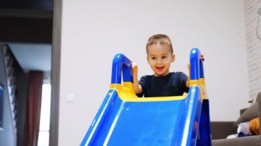 Odada duran kaydırağın arkasından sevimli erkek bebek beliriyor. Tatlı çocuk göz kırpıyor, gözlerini ovuyor ve kaydırağın tepesine tırmanıyor..