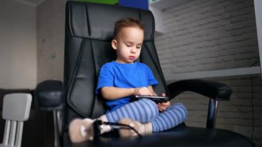 Koyu renk saçlı, mavi tişörtlü bir çocuk bacaklarını büyük bir ofis sandalyesine katlamış oturuyor. Sevimli çocuk akıllı telefon ekranına parmağıyla dokunuyor. Düşük açı görünümü.