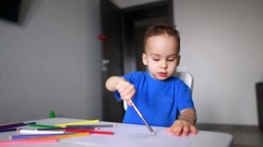 Çekici bir oğlan elinde kalemle masada oturuyor. Sevimli çocuk çiziyor ve konuşuyor. Bulanık arkaplan.
