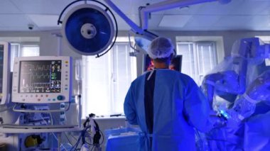 Da Vinci Robot 'un operasyon alanında mavi cüppeli bir erkek doktorun dikiz görüntüsü. Akciğer ventilatörü monitörleri ameliyat altındaki hastanın yaşam sinyallerini gösteriyor..
