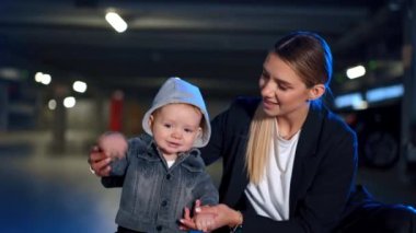 Beyaz kadın, kapüşonlu ve kot ceketli şirin bebeğini destekliyor. Anne, sevimli bir şekilde gülümseyen çocuğun elini sallıyor. Bulanık arkaplan.