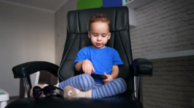 Mavi tişörtlü yakışıklı beyaz çocuk deri sandalyede oturuyor. Küçük çocuk akıllı telefonuyla oynuyor..