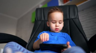 Ciddi bir Kafkasyalı çocuk, elinde telefonla dikkatle bakıyor. Çocuk büyük sandalyede oturmuş aletle oynuyor. Düşük açı görünümü.