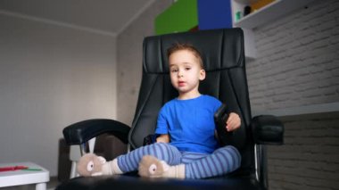 Beyaz erkek bebek büyük siyah koltukta oturuyor. Akıllı telefonu ve güneş gözlüğünü elinde tutan tatlı bir çocuk..