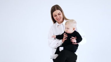 Beyaz tişörtlü mutlu bir kadın elinde siyah takım elbiseli sarışın bir bebek tutuyor. Tatlı tombul yanaklı çocuk annesinin kucağında huzur içinde oturuyor..