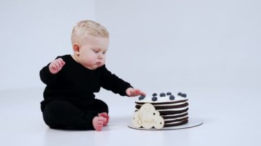 Güzel, yalınayak, siyah takım elbiseli sarışın bebek yerde oturuyor. Çocuğun yanında lezzetli bir çikolatalı pasta var ve çocuk tabelaya dokunuyor. Beyaz arkaplan.