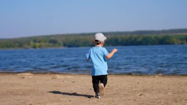 Bebeğimiz kumsaldan nehre doğru koşuyor. Enerjik çocuk taşı suya atıyor. Doğada yaz günü.