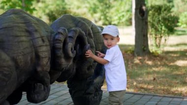 Beyaz tişört ve kep giyen güzel erkek bebek ahşap heykelin önünde duruyor. Meraklı çocuk parktaki rakamlara dokunuyor..