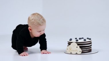 Sevimli sarışın bebek önünde duran pastaya daha da yaklaşıyor. Çocuk lezzetli tatlıya yakın. Beyaz arkaplan.