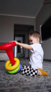 İki yaşında bir çocuk yerde oturmuş piramidi söküyor. Bebek evde oyuncaklarla oynuyor. Düşük açı. Dikey video.