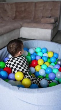 Kareli pijamalı bebek kuru havuzda yuvarlanıyor. Evde bir çocuk için aktif oyun zamanı. Dikey video.