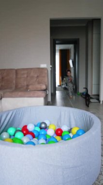 Renkli topları olan kuru bir havuz oturma odasında. Küçük çocuk odaya koştu ve havuza daldı. Kara kedi bir çocukla koşar. Dikey video.