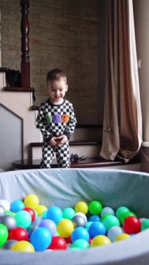 Şirin çocuk kuru havuza atlıyor. Bebek kameraya gülümseyen renkli taşakların üzerinde yatıyor. Dikey video.