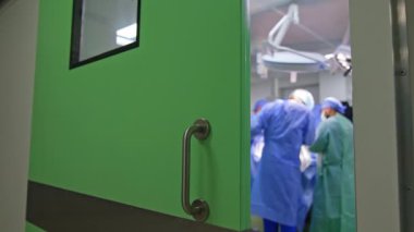Modern bir ameliyathanenin kapısını hafifçe açtı. Operasyon masasında çalışan bir grup doktor. Bulanık arkaplan.