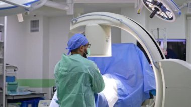 Hastanın üstündeki C-Arm makinesinin başında duran sakallı bir doktorun dikiz aynasından. Başka bir cerrah ameliyat masasına gelir..