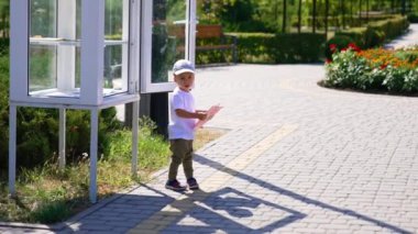 Şapkalı sevimli bebek kitap değişimi için standda duruyor. Yazın parkta elinde kitap tutan bir çocuk..
