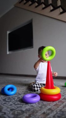 Beyaz tişörtlü ve kareli pantolonlu çocuk oyuncağına doğru koşuyor. Çocuk evde bir piramit inşa ediyor..