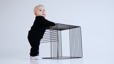 Siyah takım elbiseli sarışın bir bebek ve beyaz çoraplar metal tel küpün yanında duruyor. Çocuk elini küpten tutup gülümsüyor. Düşük açı. Beyaz arkaplan.